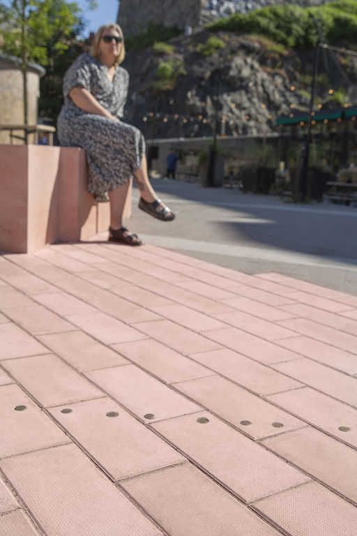 En rosa plattlagd yta av våtpressad betong ligger under ett monument på Esperantoplatsen i Göteborg. En kvinna sitter i bakgrunden på kanten på monumentet.