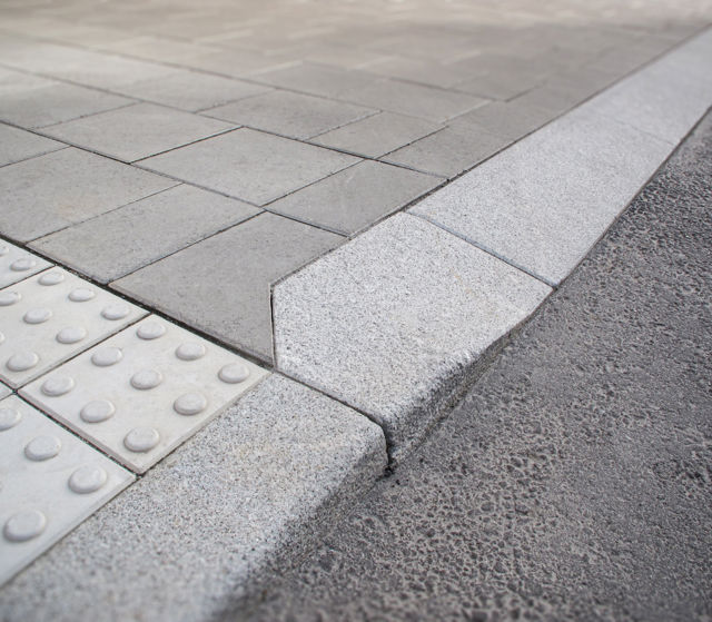 Portstens i natursten;kompletterad med taktila betongplattor med orienteringsmönster samt släta betongplattor i gångyta – i övergång mellan gångbana och körbana.
