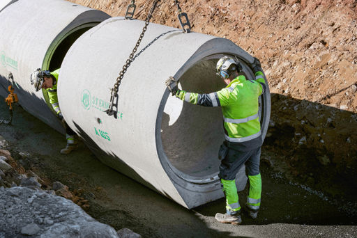Två anläggningsarbetare installerar ett stort fotrör från S:t Eriks i ett schakt;med hjälp av kran och lyftkedjor. Personalen bär skyddsutrustning och arbetar i en säker miljö vid markarbete för dagvatten eller avlopp.