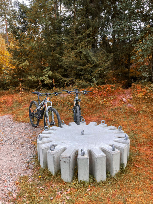 Cykelstället Kuggen på plats i ett skogsbryn i höstfärger med två mountainbikes parkerade i stället.