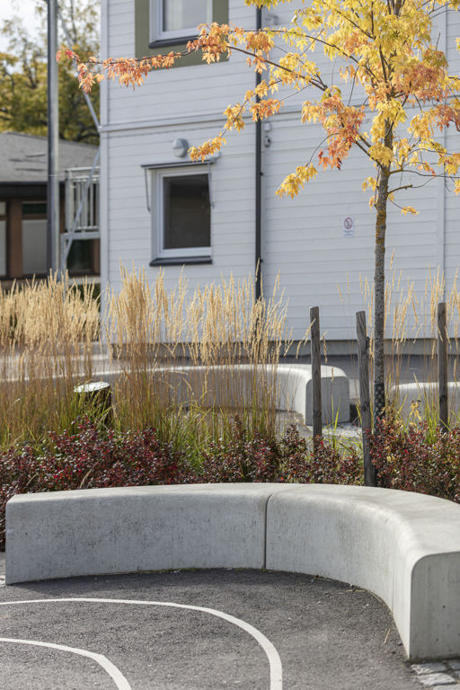 Flera Outline-bänkar bildar en ringlande orm på en skolgård med gula höstträd och vitt hus i bakgrunden