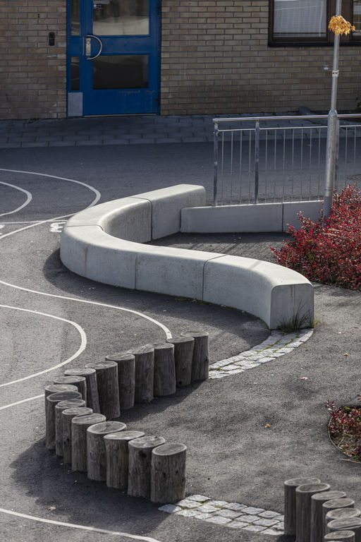 Flera Outline-bänkar bildar en ringlande orm samt trästubbar i en linjepå en skolgård med röda buskar