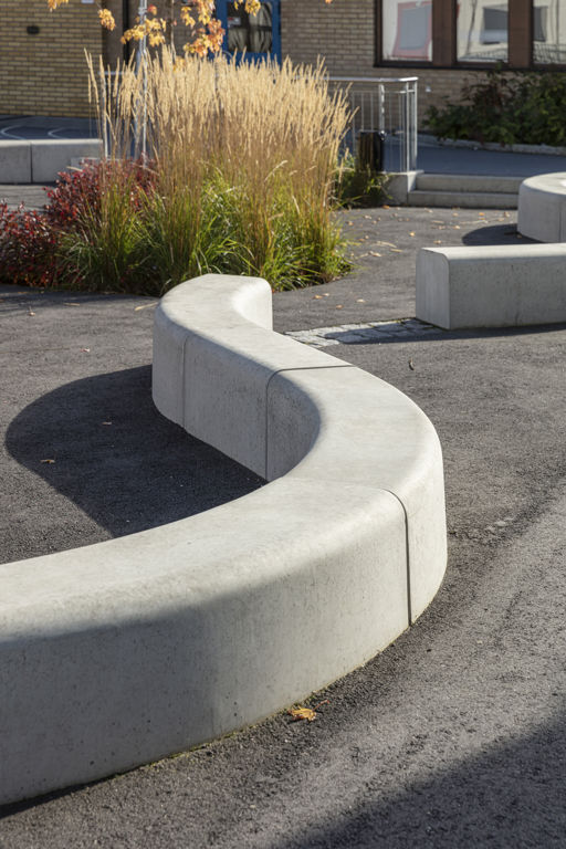 Flera Outline-bänkar bildar en ringlande orm på en skolgård med gul vass och gröna buskar